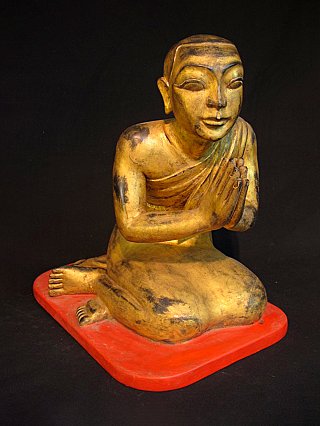Wooden monk statue with hands in Namaskara mudra, seated on red base, angled front view