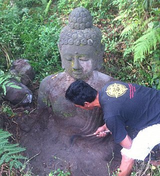 Große sitzende Buddha-Statue aus Lavastein aus Indonesien, halb eingegraben im Grün, Mann reinigt die Oberfläche